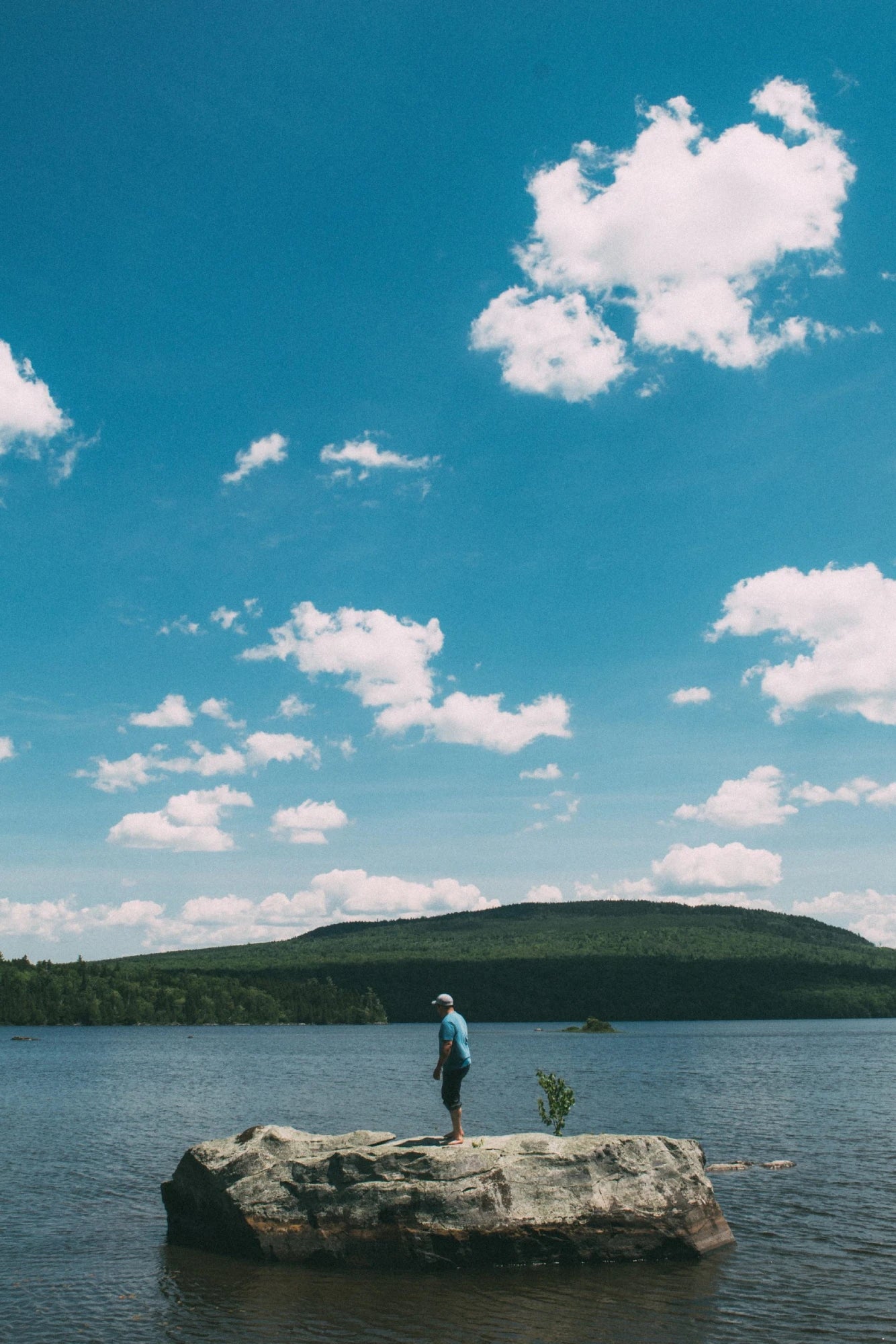 A lone man stands on a rock in the middle of a river on a sunny day.